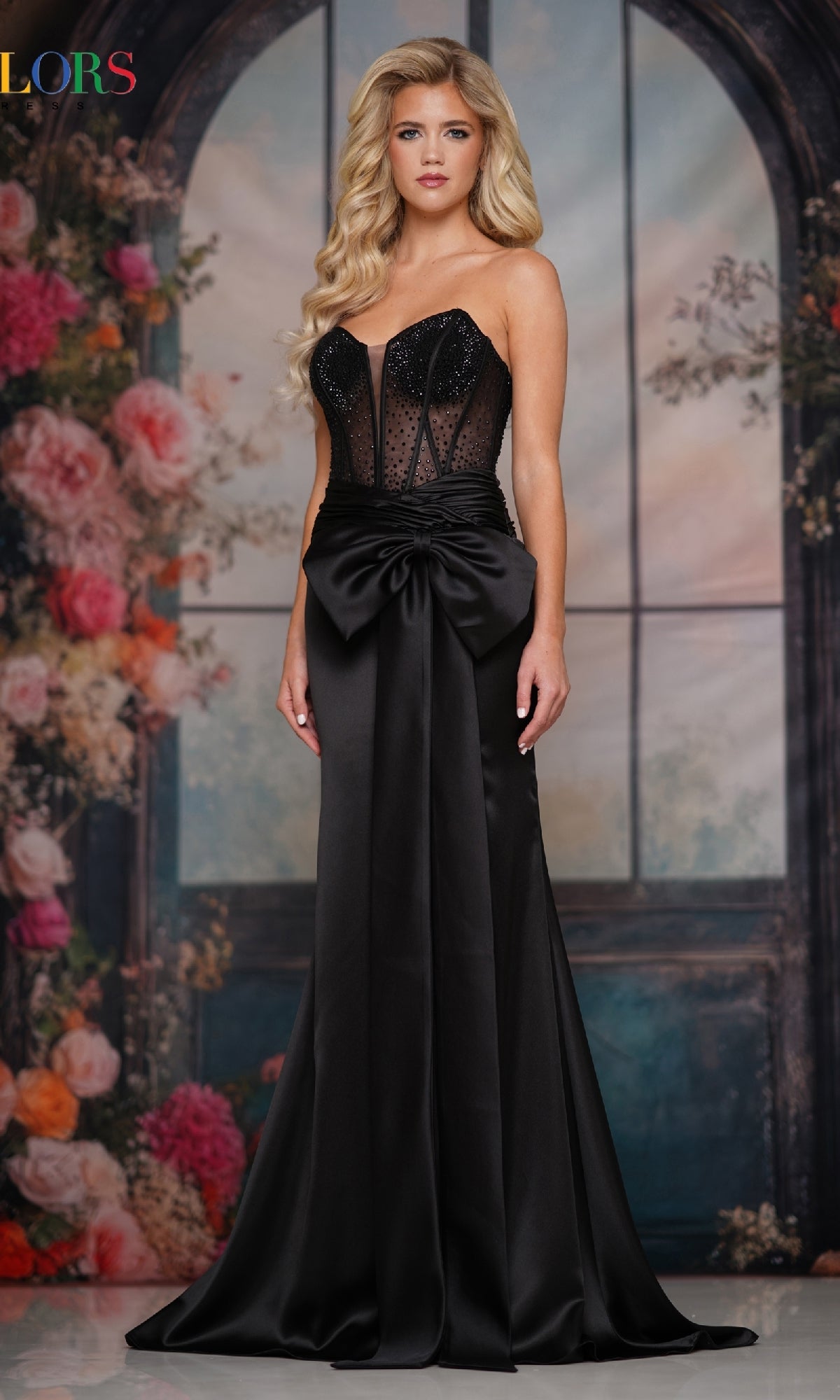 A woman wearing the Long Prom Dress: Colors Dress 3769, with a strapless rhinestone corset bodice and satin bow, stands before a floral and arched window backdrop.